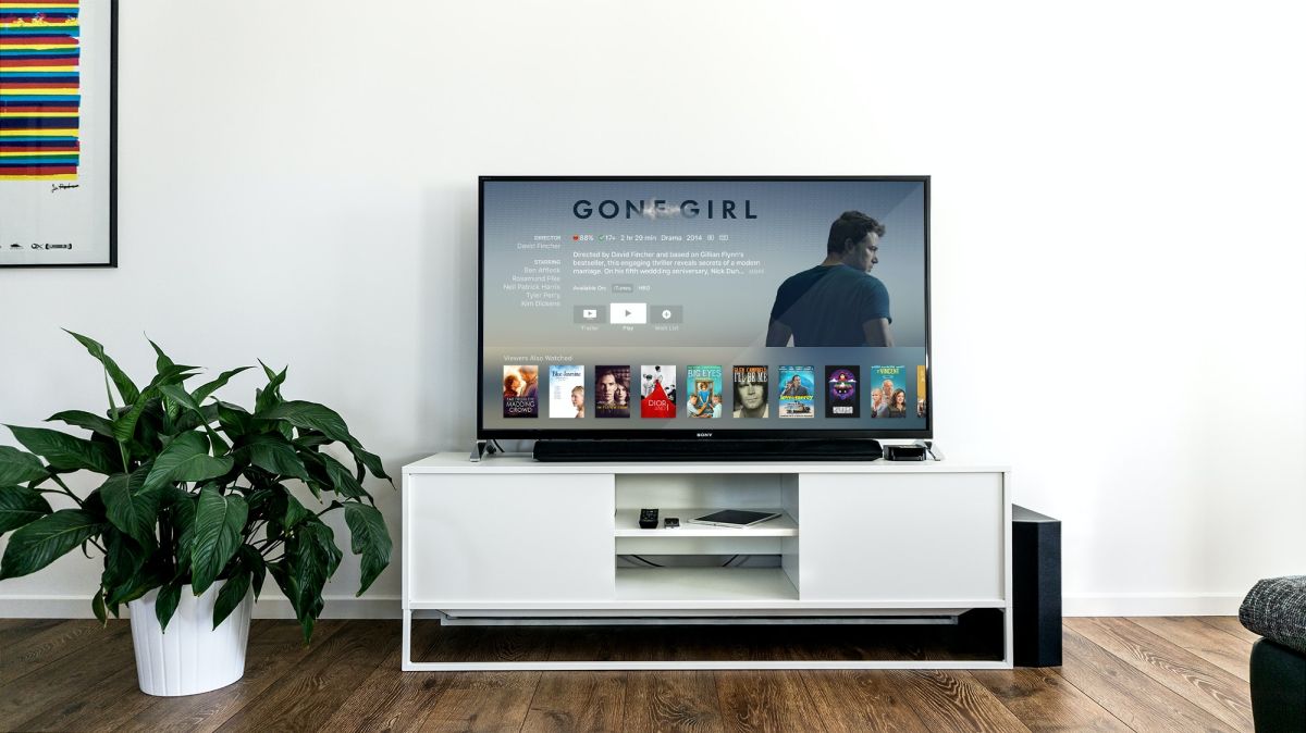 A television with soundbar sitting on a white entertainment center with a white background and a large potted plant to the left.